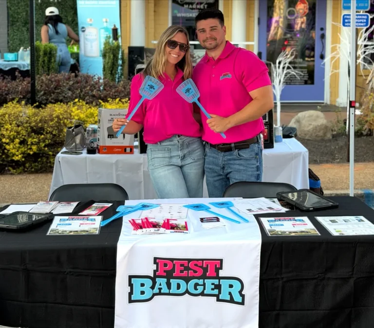 A smiling man and woman in matching bright pink "Pest Badger" polo shirts standing behind a promotional table, holding up blue fly swatters.